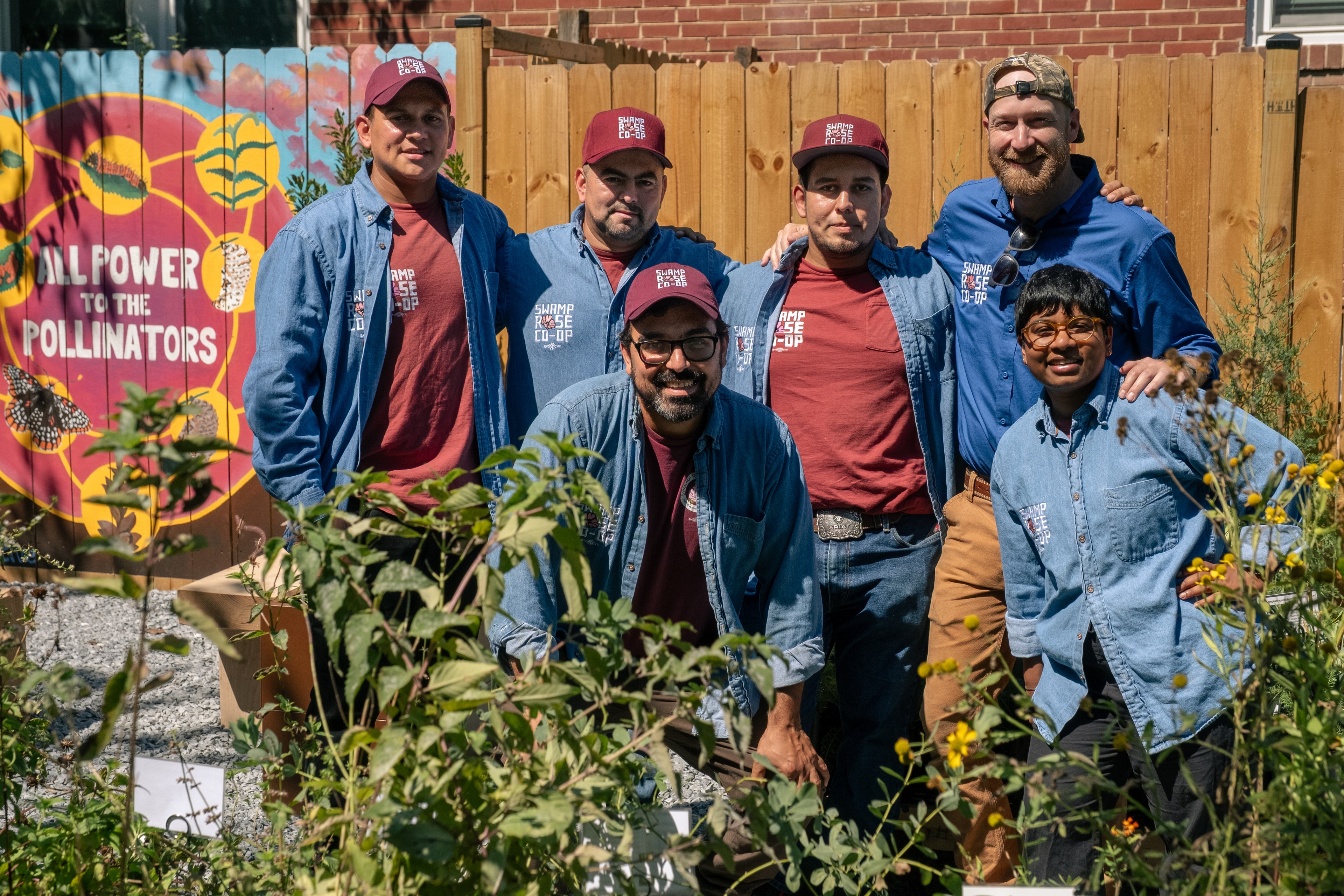 Workers pose in a garden, wearing blue work shirts reading "Swamp Rose Co-op" and standing in front a bright hand-painted sign that says "All Power to the Pollinators"